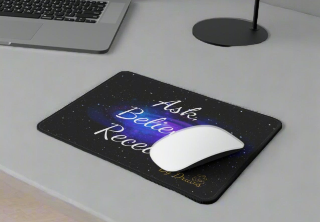 Close-up of an Ask Believe Receive mouse pad on a desk with a laptop and coffee mug.