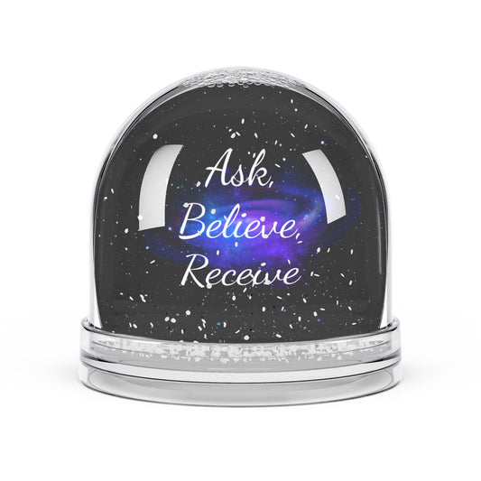 Clear snow globe with a black galaxy background and the white affirmation text “Ask. Believe. Receive.” surrounded by floating snowflakes