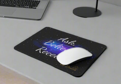 Close-up of an Ask Believe Receive mouse pad on a desk with a laptop and coffee mug.